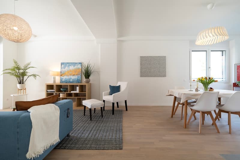 Open living and dining area with wooden floor, white walls, blue sofa, white chairs, wooden table, dining chairs, wooden shelf, plants, lamps, and artwork.
