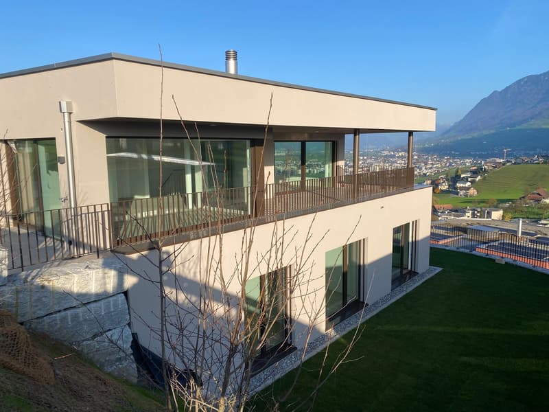 Modern architectural design, light-colored exterior, large glass windows, balcony with metal railings, elevated terrain, lush green grass.