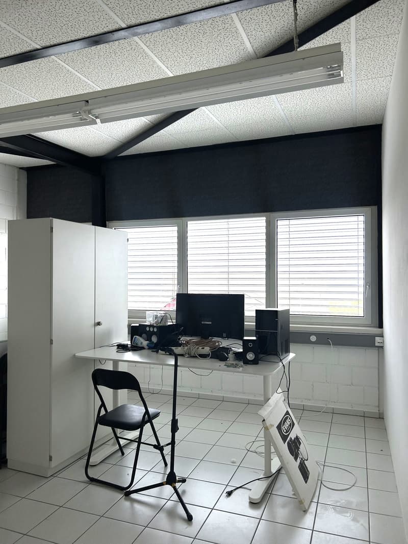 white tiled floor, white ceiling with fluorescent lighting, white office desk, black folding chair, computer monitor, speakers, storage cabinet