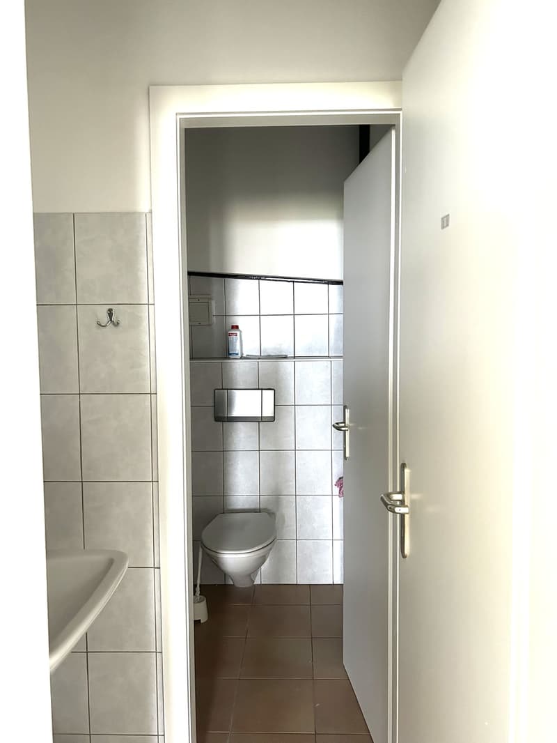 A small bathroom with white walls, brown tiled floor, white toilet, a sink, and open door.