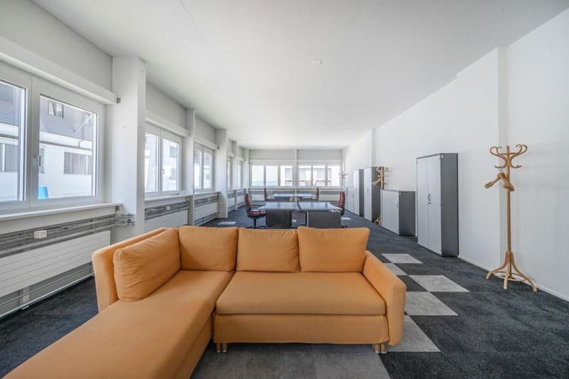 Spacious room with modern decor, white walls, grey floor, orange sectional sofa, wooden coat rack, several desks and office chairs, large windows, storage cabinets