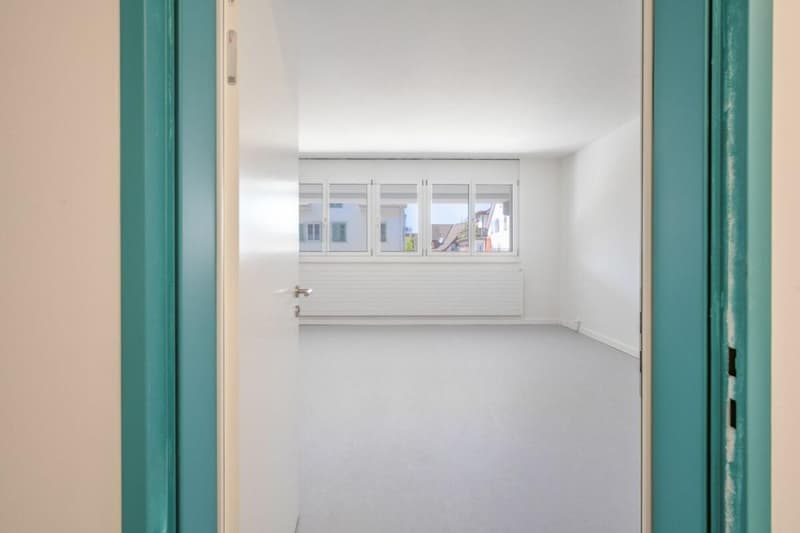 empty room, white walls, white floor, windows with views of outside buildings, radiator