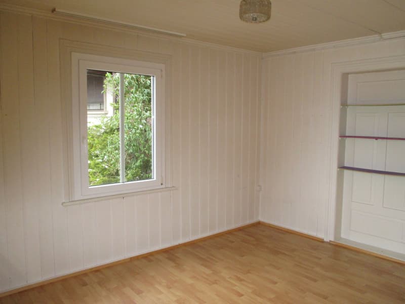 Empty room with white walls, wooden floor, window overlooking outside.