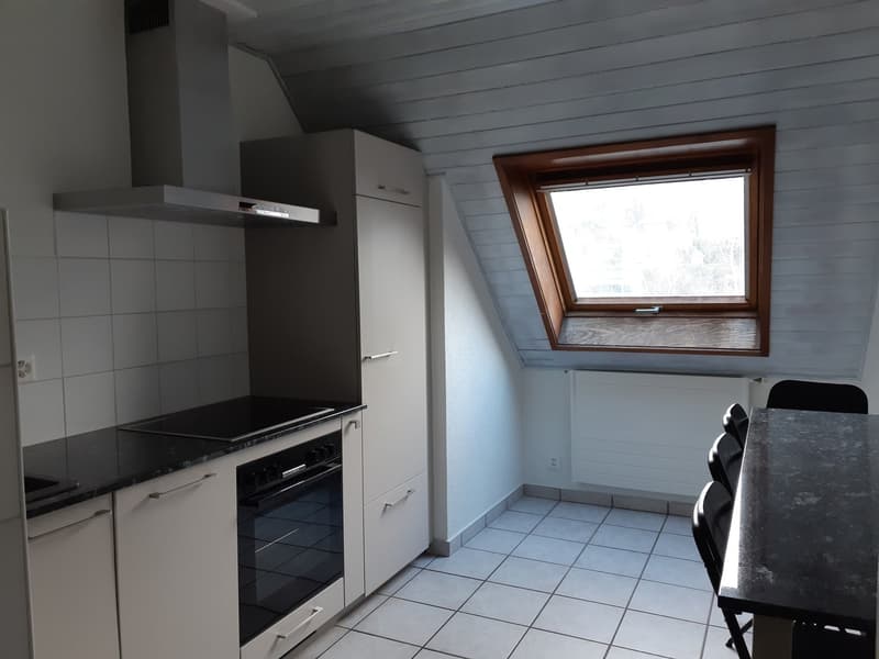 The image shows a kitchen area with a built-in oven, a dishwasher, and a window with a wooden frame. The walls are tiled, and the floor is covered with square tiles. There is a dining table with chairs in the corner of the room.