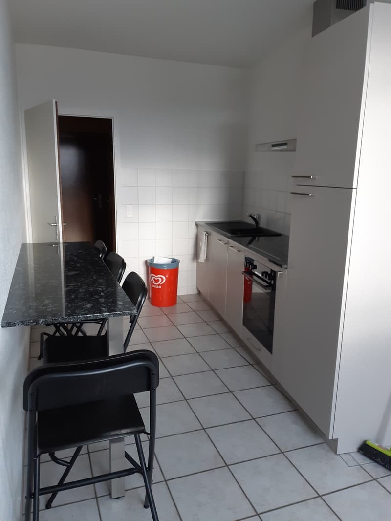 The image shows a kitchen area with a granite countertop, a sink, a stove, and a refrigerator. There is also a small dining table with black chairs. The floor is tiled, and the walls are white. A red trash can is visible in the corner.