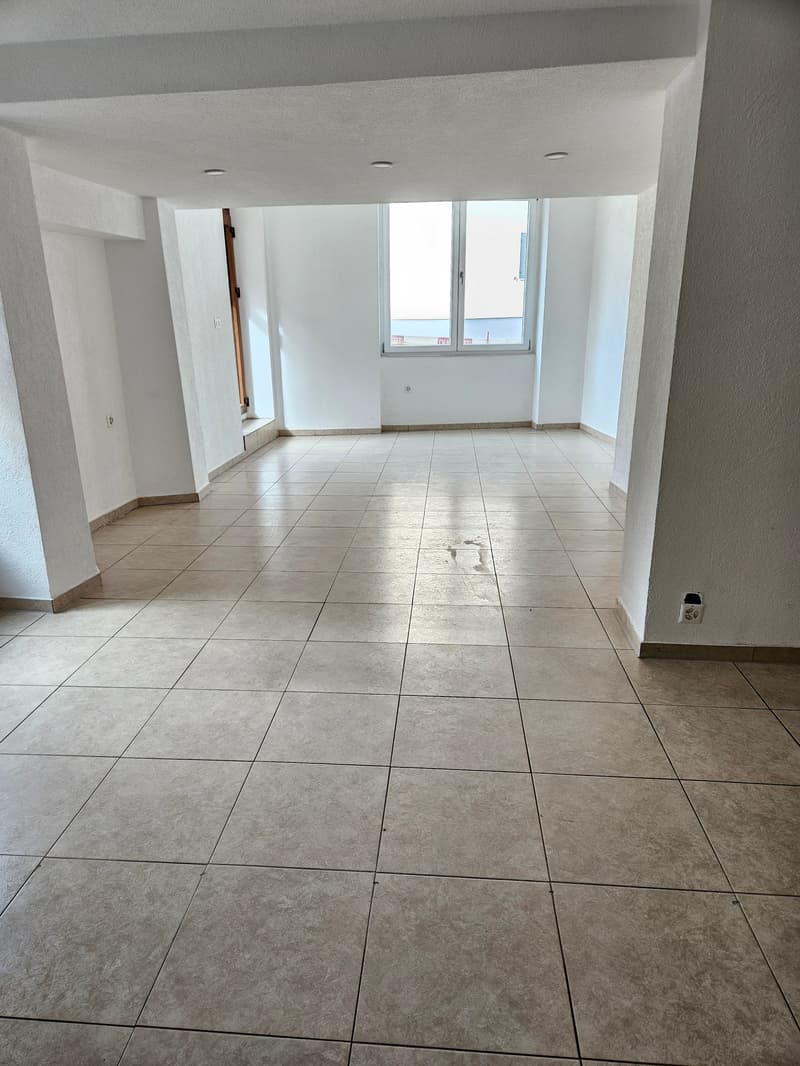 The image shows a spacious room with white walls and a tiled floor. The room has large windows that provide natural light. The space appears to be empty, with no furniture or decor visible.