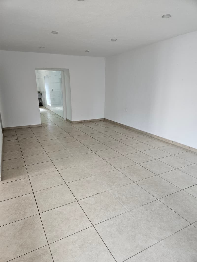 Large open room with white walls and gray tile flooring. The room appears to be empty, with no visible furniture or fixtures. There is an archway leading to another room or area visible in the background.