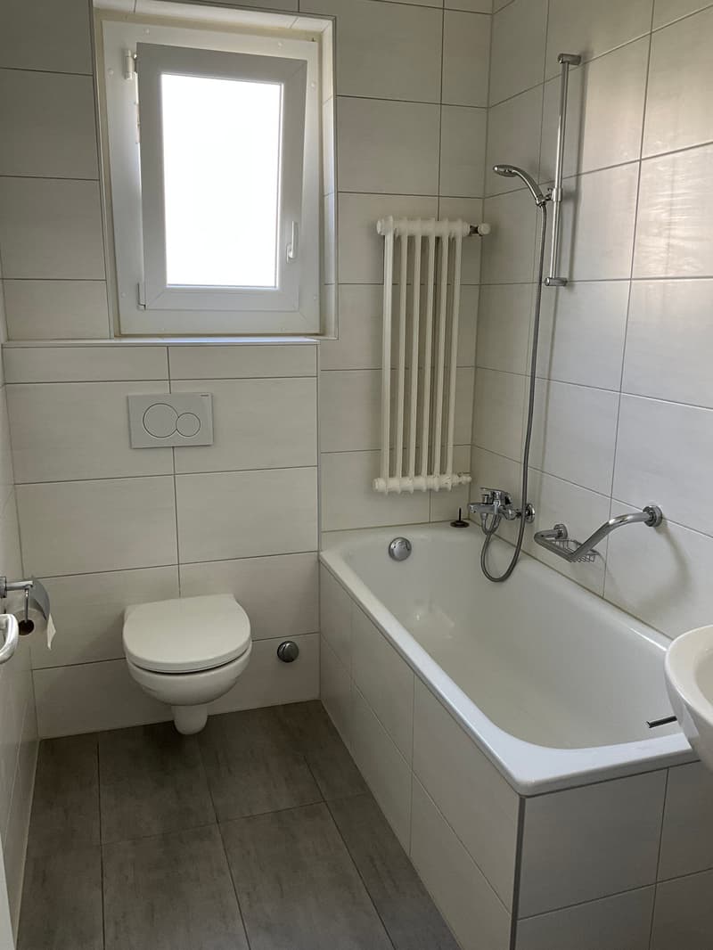 Bathroom with white tiles, bathtub, toilet, sink, window, radiator, shower head, and towel rack.