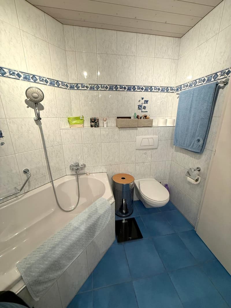Bathroom with bathtub, toilet, shelves, towel rack, blue tiled floor, white walls, shower head and towels.