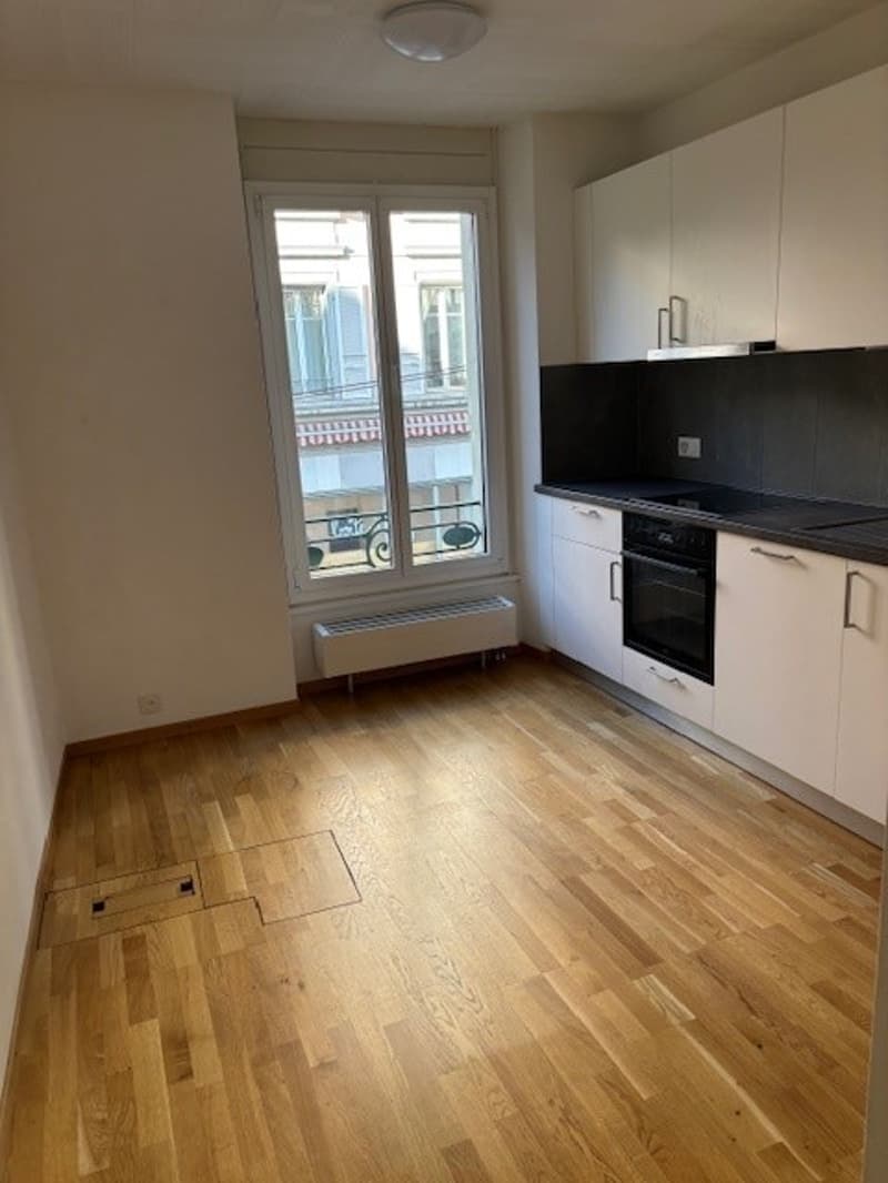 Hardwood floor, white kitchen cabinets, a white window, a gas range and oven in the corner, a heater under the window, a light on the ceiling