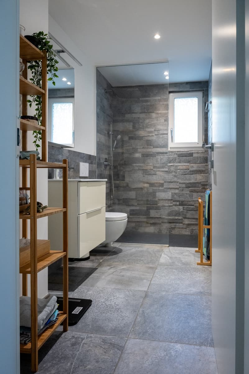 Bathroom with white toilet, white cabinet, wooden shelves, gray tiles, glass shower enclosure, and a window.
