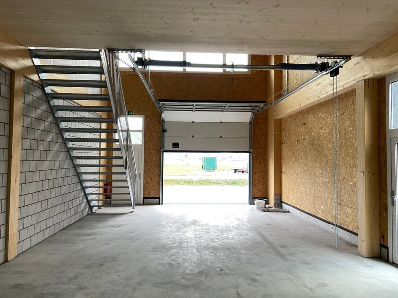 An empty garage with white tile walls, wooden beams, a large door, and a staircase.