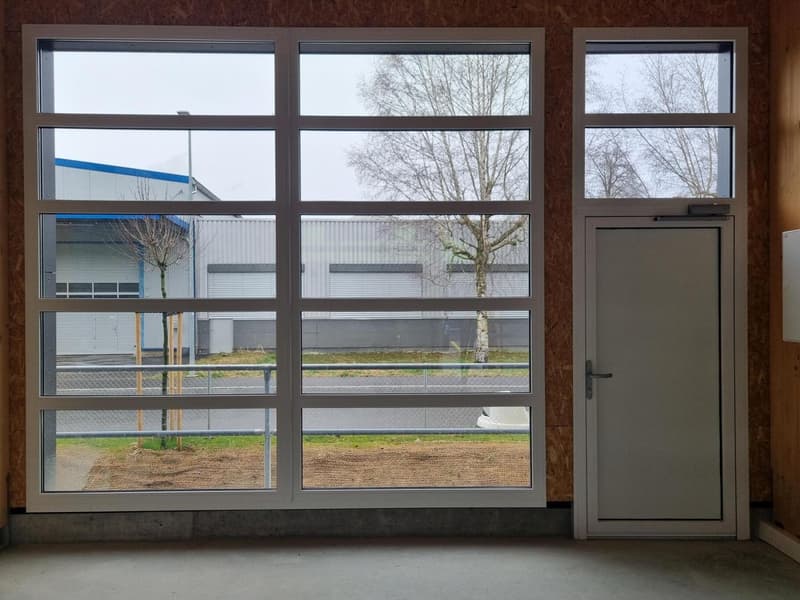 The image shows a commercial building with large windows overlooking a parking area and some trees outside. The building has a gray exterior and the windows have a modern, industrial-style design. There appears to be a door on the right side of the buildi