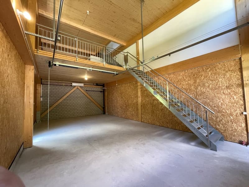 The image shows a spacious garage or storage area with a wooden ceiling, concrete floor, and a metal staircase leading to an upper level. The space appears to be unfinished, with exposed wooden beams and OSB board walls.