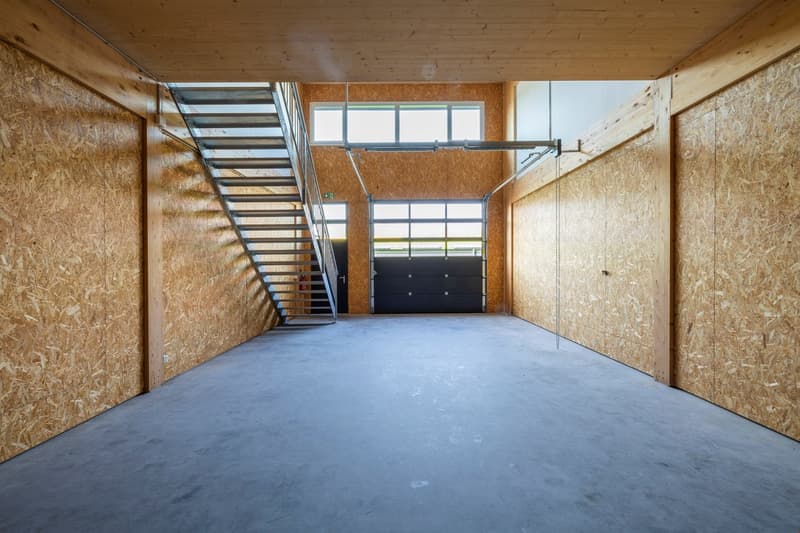 Large open space with concrete floor, wooden walls, and a metal staircase. The space appears to be a garage or storage area with a garage door visible in the background.