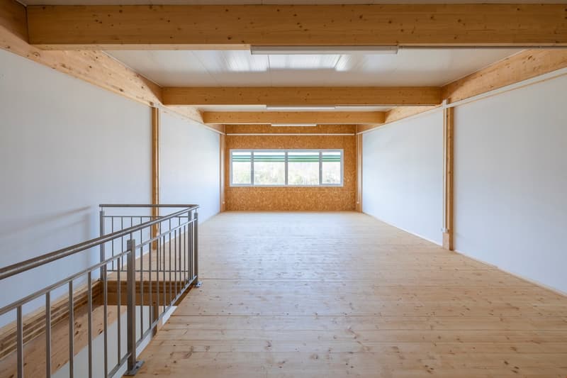 Large open space with wooden beams and ceiling, white walls, and hardwood flooring. The room has a loft-like feel with a metal railing overlooking the main floor area.