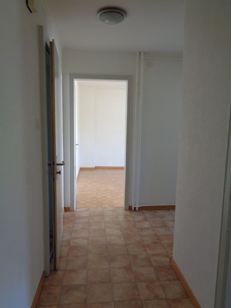 The image shows a hallway with white walls and a tiled floor. The hallway leads to another room, which can be seen through the open doorway.