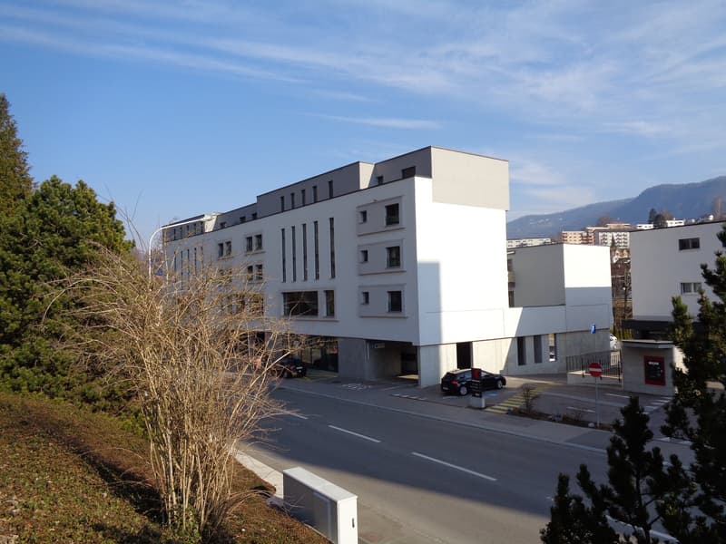 A modern, multi-story building with a white exterior and dark accents. The building has several windows and appears to be located near a road with a parked car visible.