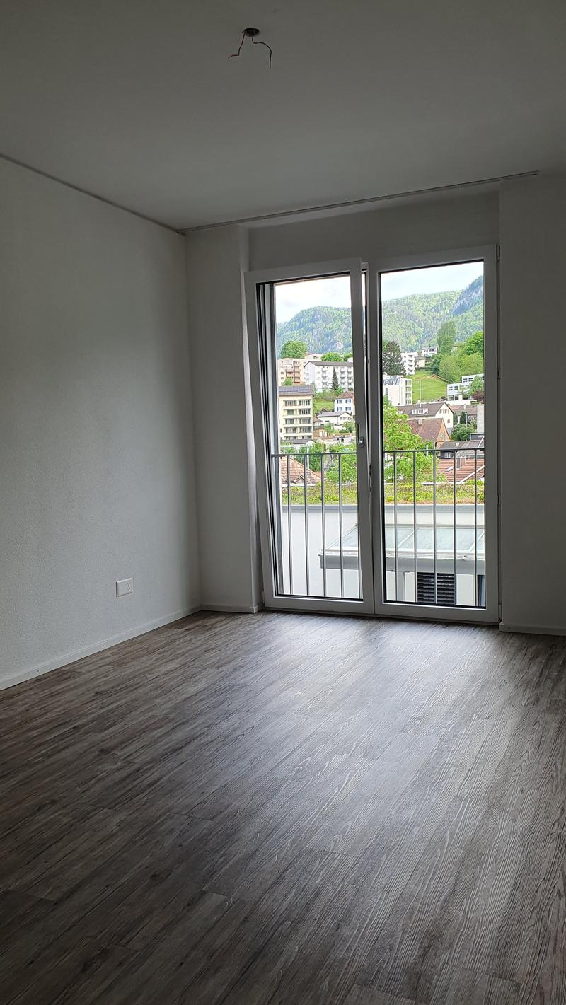Spacious bedroom with large windows offering a scenic view of the surrounding buildings and landscape. The room has hardwood-style flooring and white walls.