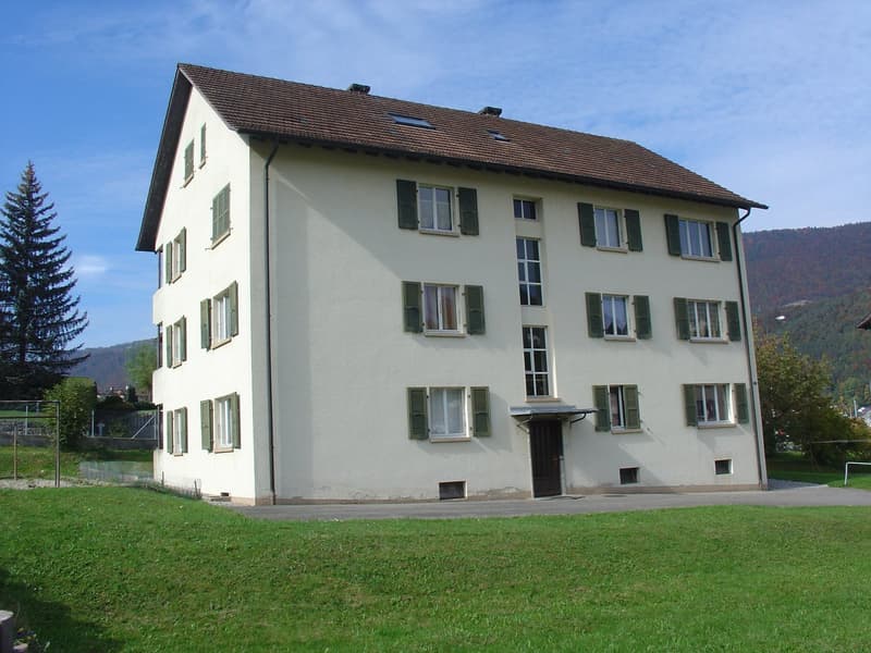 White multi-story building with multiple windows and green shutters, brown roof, entrance, surrounded by lawn