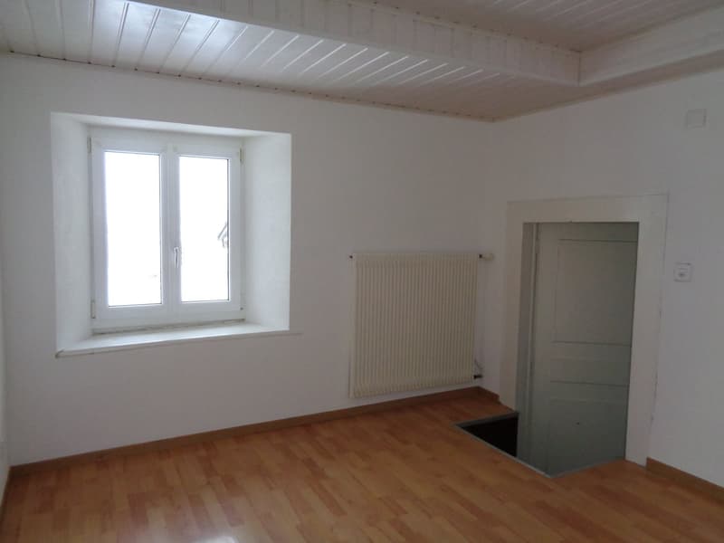 An empty room with white walls, wooden floor, a window with two glass panes, a radiator, a small hidden entrance, and a door.