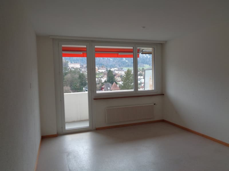 Empty room with sliding glass door leading to balcony, white walls, large window, white ceiling, wooden floor, white heater below window