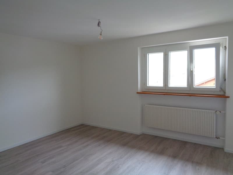 An empty room with wooden floors, white walls, a white radiator, and a large window with a wooden sill.