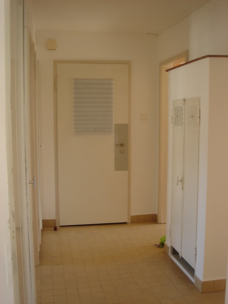 Empty hallway with white walls, tiled floor, white door, and a locker.