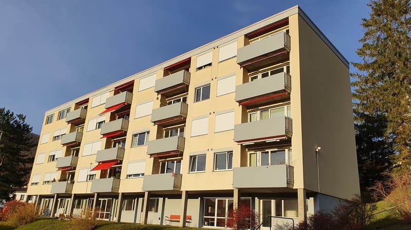 Multi-story apartment building, beige exterior, multiple balconies, red awnings, glass windows, entrance with glass doors, surrounded by greenery