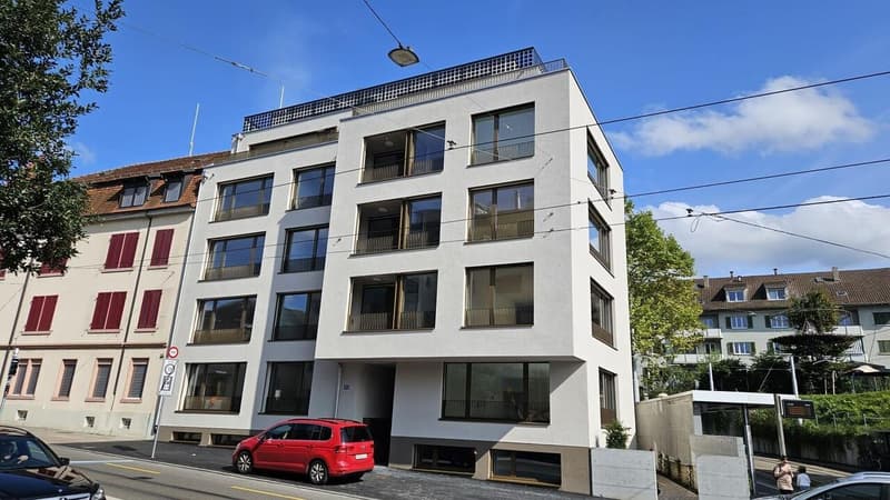 white modern building with 4 floors, multiple windows, solar panels on the roof, street light, no parking place visible, small trees, bus stop
