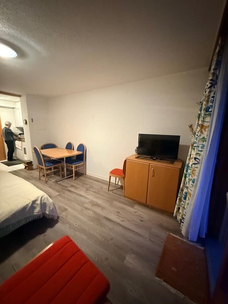 small room, single bed, table with four chairs, red bench, television, person in doorway