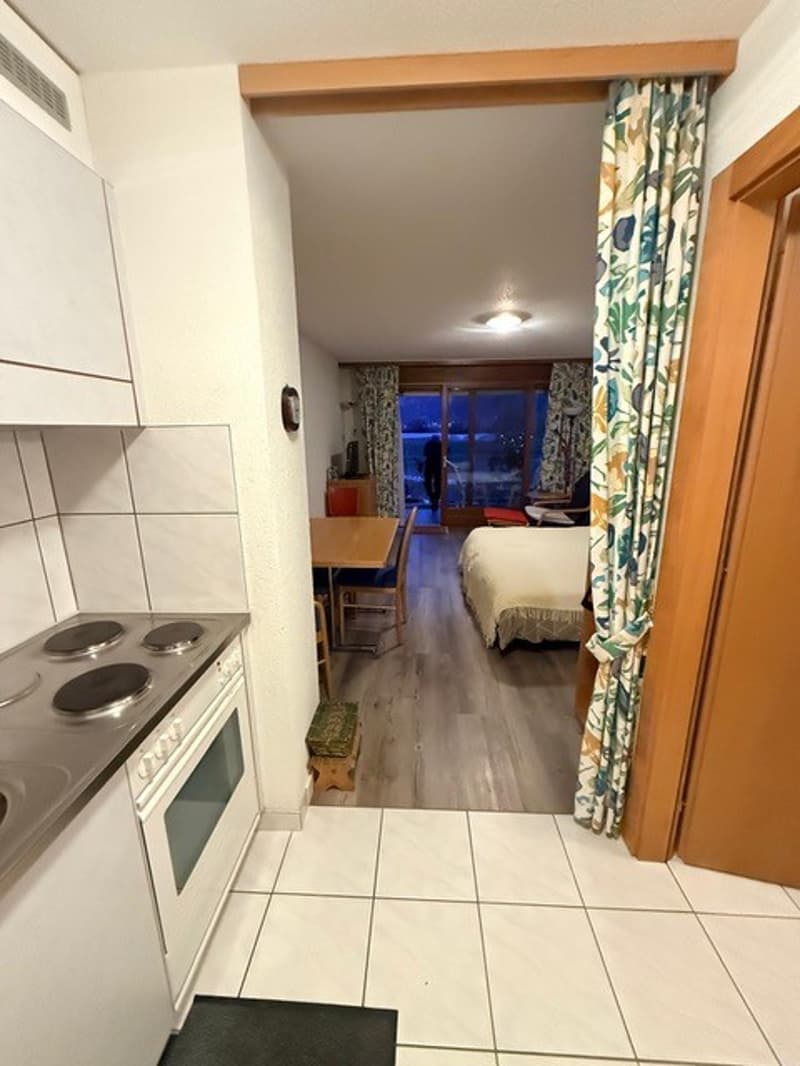 White kitchen with a gas stove, an oven, white cabinets, and a dining area with a table and chairs. There's a corridor leading to a bedroom with a bed and a small rug.