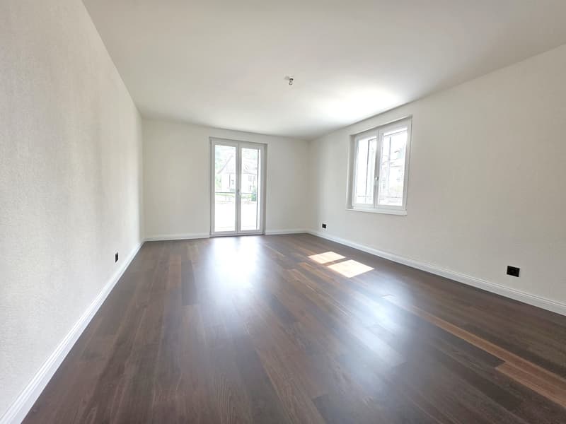 Spacious living room with hardwood flooring, white walls, and large windows providing ample natural light. The room has a clean and minimalist design aesthetic.