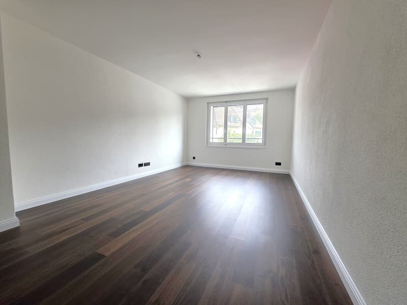 Spacious room with white walls, hardwood flooring, and large windows providing natural light. The room appears to be empty, ready for furnishing and decorating.
