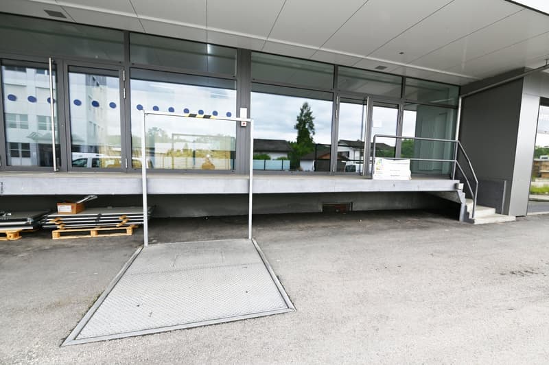 concrete elevated ground floor with glass walls and railings, a wheelchair ramp, stairs, and a parking area
