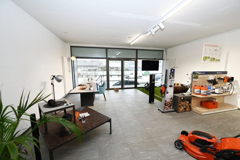 The image shows a commercial space or showroom with large windows, a tiled floor, and various tools and equipment displayed. There is a desk and seating area, as well as a lawn mower and other outdoor equipment visible, suggesting this may be a home impro