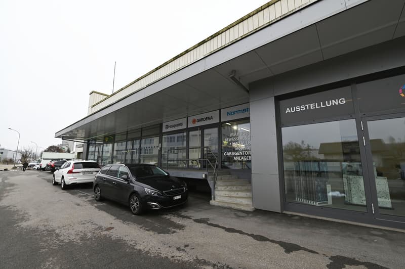 2-story commercial building with signage for Normstahl, Husqvarna, and Gardena brands. The building has a large glass facade and a covered entrance area. There are several parked cars in the front of the building.