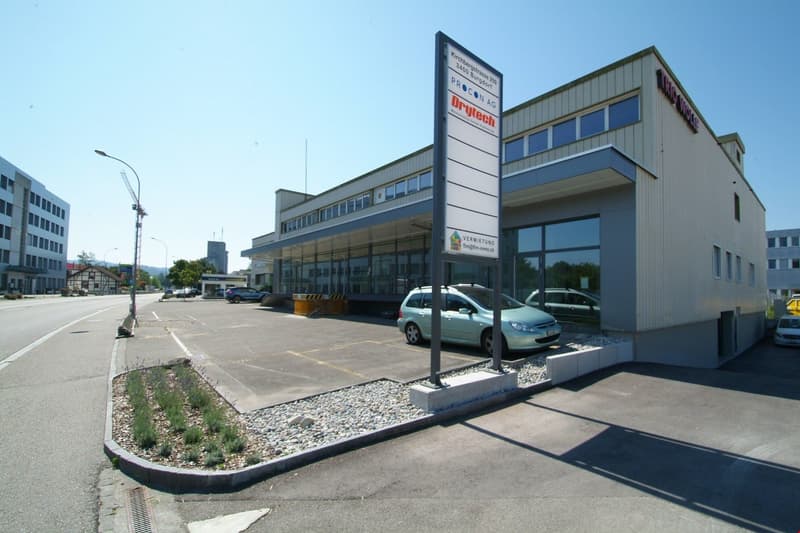 2-story commercial building with Procon AG branding, large windows, parking lot in front, landscaping with rocks and plants