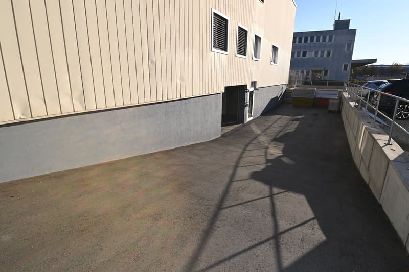 Wide driveway with paved surface, concrete walls, metal railings, and adjacent building with white panels and windows.