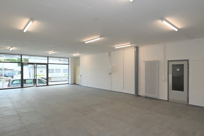 Empty room with white walls, gray floor, large windows, and ceiling lights