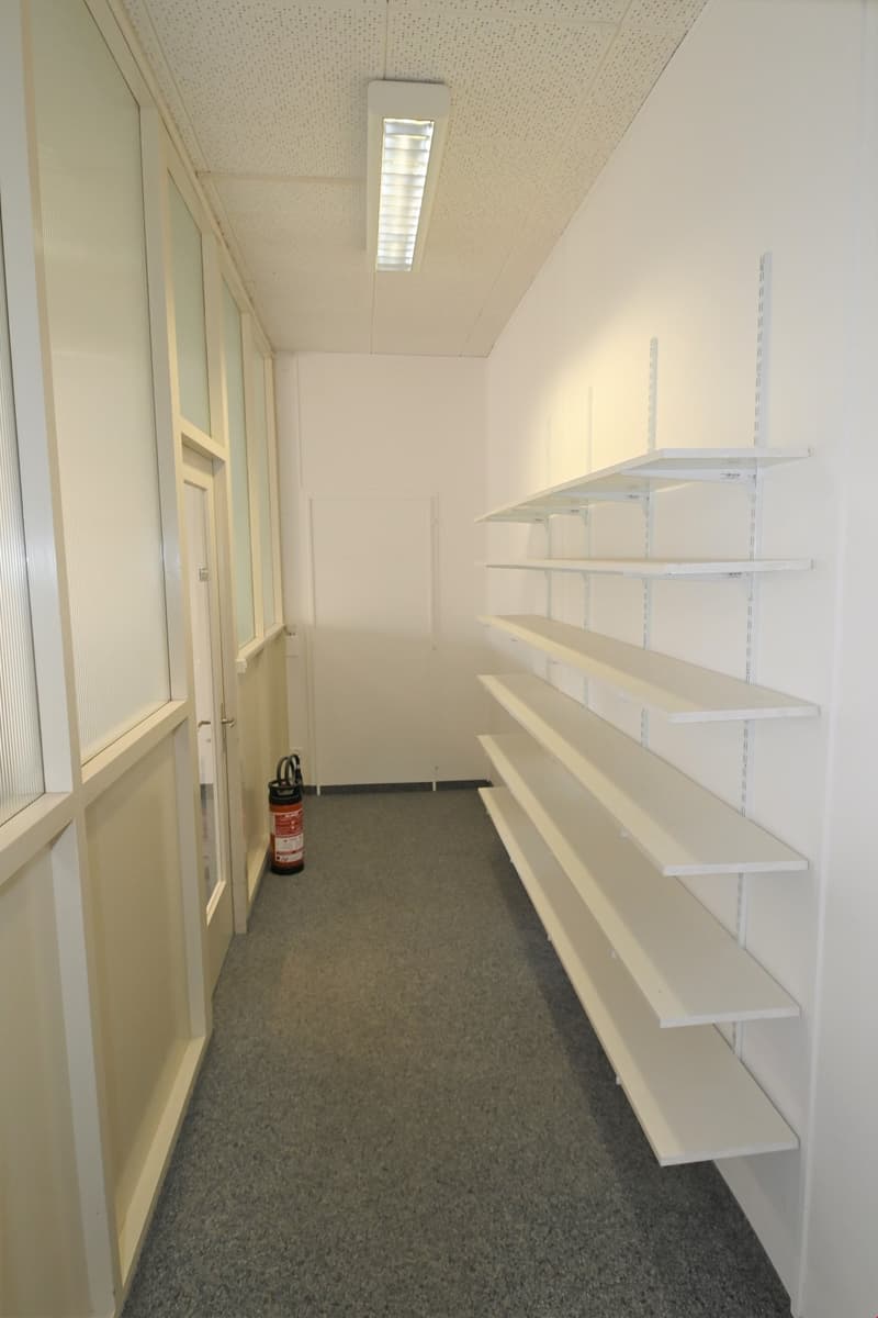 Empty room with white walls, gray floor, white shelves, and a fire extinguisher.