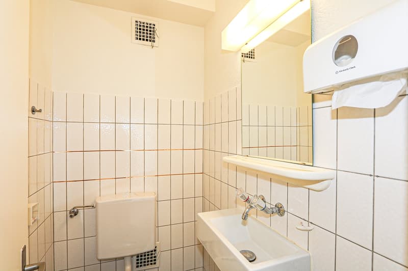white tiled bathroom with sink, toilet, mirror, and towel dispenser