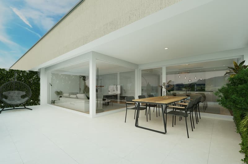 Modern open-plan living space with large glass windows and doors leading to a balcony or terrace. The space includes a dining area with a wooden table and chairs, and a living room with comfortable seating. The exterior walls are white and the flooring is