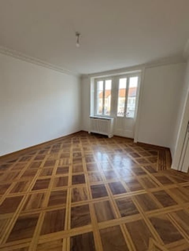 Empty room with wooden parquet flooring, white walls, large window, radiator