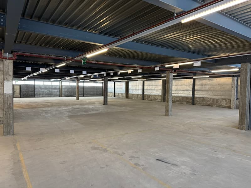 Open parking area with concrete floor, metal beams, and exposed ceiling. Large empty space with yellow markings on the floor.
