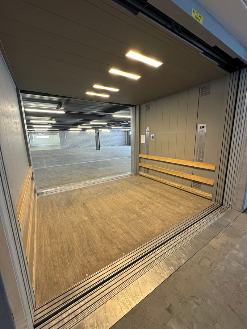 An open garage space with a concrete floor, ceiling-mounted lighting, and sliding doors. The interior has a minimalistic design with a wall and an elevator visible on the right.