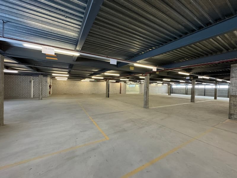 Large empty parking lot with yellow painted lines, grey concrete floor, metal beams on ceiling, exposed brick wall, emergency extinguisher, and fluorescent lighting.
