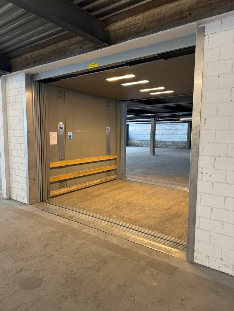The entrance of an underground parking facility, the entrance is an opening to a car lift. The ground floor is raised. The space is empty.