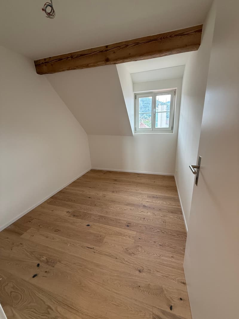 This image shows a bedroom with a wooden beam running across the ceiling. The room has a hardwood floor and a small window that provides natural light. The walls are painted a light, neutral color.