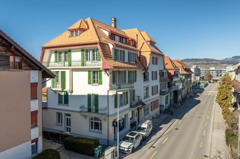 This image shows a street in a European town, with colorful multi-story buildings lining the road. The buildings have tiled roofs and balconies, and there are cars parked along the street. The sky is a bright blue, indicating a sunny day.